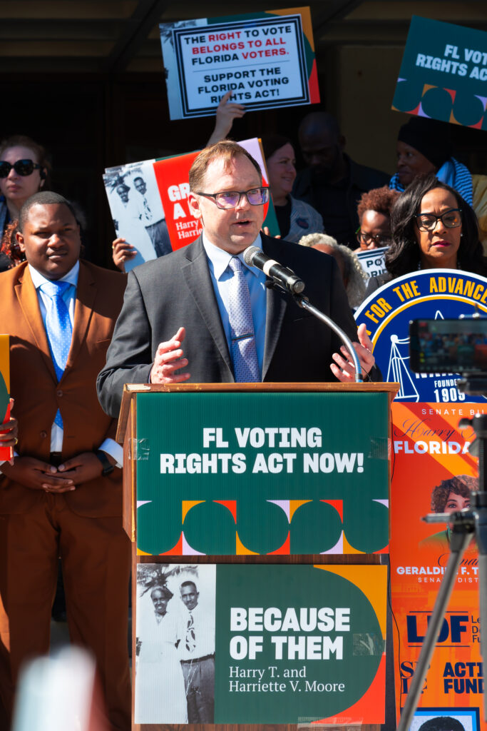 An All Voting is Local Action team member standing at a lectern with a sign that says "FL Voting Rights Act Now!" on it.