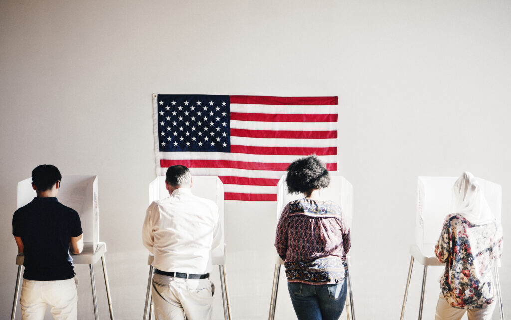 People standing at polling booths with an American flag on the wall in front of them.